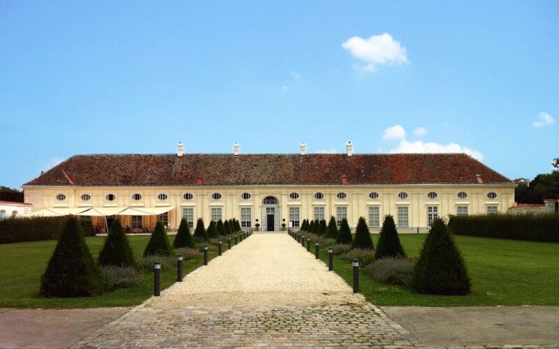 Ein langgestrecktes, einstöckiges Gebäude mit einem Ziegeldach und mehreren Fenstern steht am Ende eines Kiesweges, der von beschnittenen Büschen und Lampen gesäumt ist, unter einem klaren blauen Himmel.