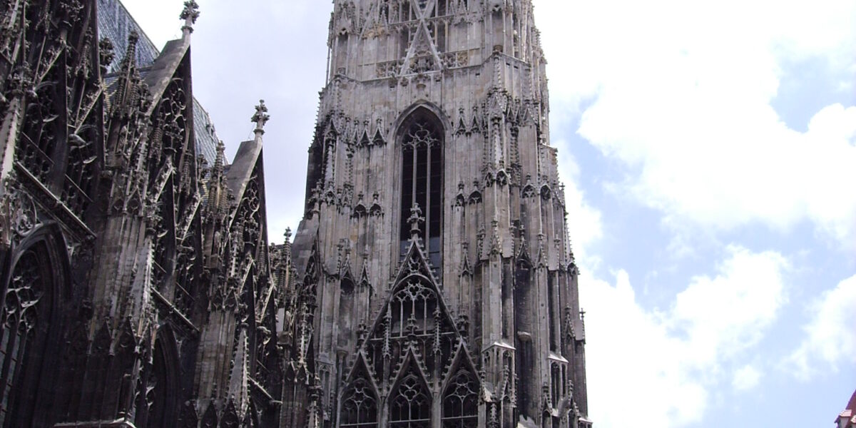 Hohe gotische Kathedrale mit kunstvollen Steindetails und einer zentralen Turmspitze, umgeben von historischen Gebäuden unter einem teilweise bewölkten Himmel.