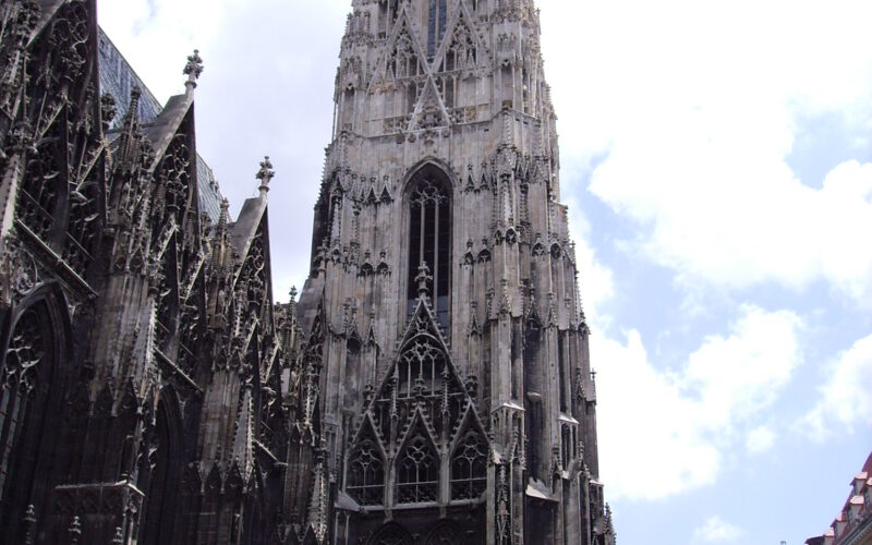 Hohe gotische Kathedrale mit kunstvollen Steindetails und einer zentralen Turmspitze, umgeben von historischen Gebäuden unter einem teilweise bewölkten Himmel.