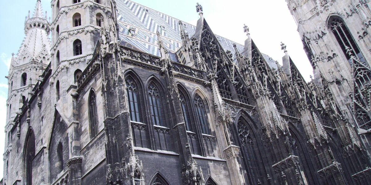 Kathedrale im gotischen Stil mit Spitzbögen, detaillierten Steinmetzarbeiten, hohen Türmen und einem gemusterten Ziegeldach unter einem teilweise bewölkten Himmel.