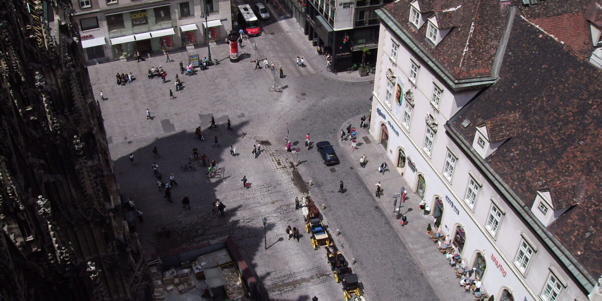 Luftaufnahme eines kopfsteingepflasterten Stadtplatzes mit Fußgängern, geparkten Autos und umliegenden mehrstöckigen Gebäuden an einem sonnigen Tag.