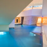 Hallenbad unter einer modernen architektonischen Struktur mit umlaufenden Wegen, Steinmauern und beleuchtetem Sitzbereich im Hintergrund.