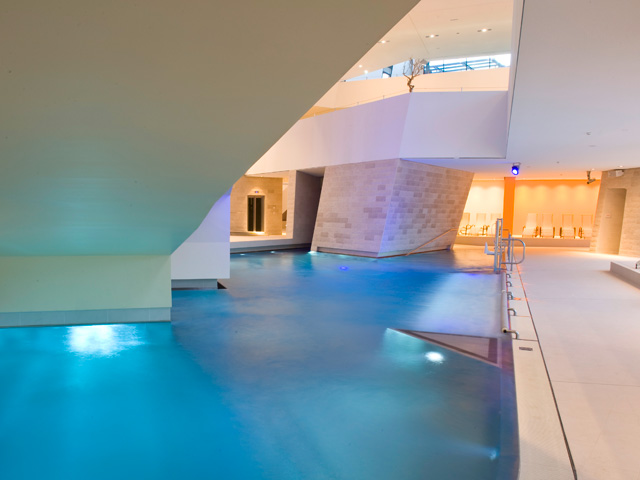 Hallenbad unter einer modernen architektonischen Struktur mit umlaufenden Wegen, Steinmauern und beleuchtetem Sitzbereich im Hintergrund.