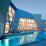 Modernes Gebäude mit großen, eckigen Glasfenstern und einem in der Abenddämmerung beleuchteten Außenschwimmbad. Rund um den Pool sind Liegestühle aufgestellt.