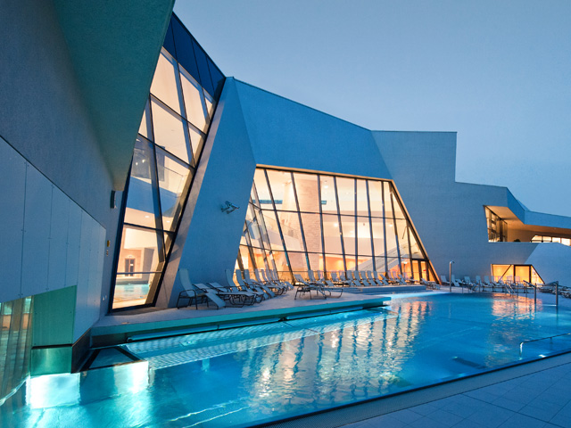 Modernes Gebäude mit großen, eckigen Glasfenstern und einem in der Abenddämmerung beleuchteten Außenschwimmbad. Rund um den Pool sind Liegestühle aufgestellt.