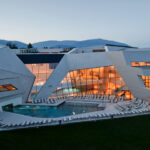 Modernes Gebäude mit kantiger weißer Architektur, großen Glasfenstern und einem von Liegestühlen umgebenen Außenpool; im Hintergrund sind die Berge zu sehen.