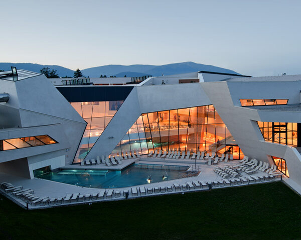 Modernes Gebäude mit kantiger weißer Architektur, großen Glasfenstern und einem von Liegestühlen umgebenen Außenpool; im Hintergrund sind die Berge zu sehen.