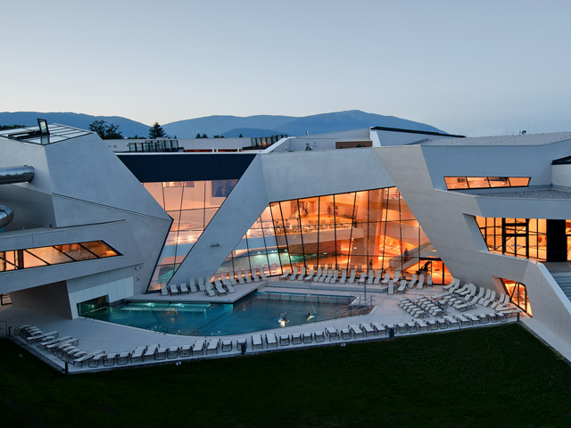 Modernes Gebäude mit kantiger weißer Architektur, großen Glasfenstern und einem von Liegestühlen umgebenen Außenpool; im Hintergrund sind die Berge zu sehen.