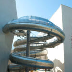 Eine große, spiralförmige Metallrutsche ist an der Außenseite eines modernen, weißen Gebäudes angebracht, an dessen Wand in bunten Buchstaben das Wort "KARAHOF" steht.