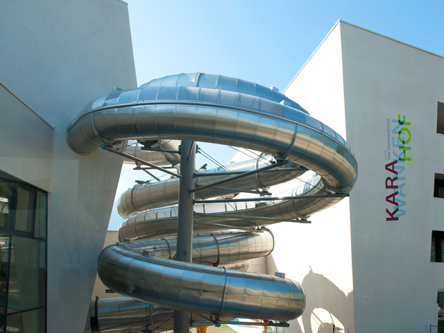 Eine große, spiralförmige Metallrutsche ist an der Außenseite eines modernen, weißen Gebäudes angebracht, an dessen Wand in bunten Buchstaben das Wort "KARAHOF" steht.