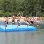 Sechs Jungen in Badehosen springen von einem blauen Schwimmsteg in einen See, im Hintergrund sind Bäume und ein Sandstrand zu sehen.