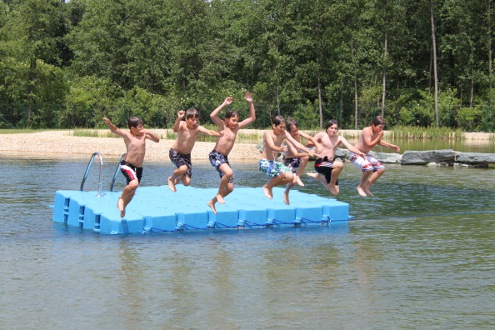 Sechs Jungen in Badehosen springen von einem blauen Schwimmsteg in einen See, im Hintergrund sind Bäume und ein Sandstrand zu sehen.