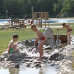 Drei Jungen in Badeanzügen spielen mit Wasser und Steinen in einem flachen Bach in einem Park im Freien, wobei im Hintergrund andere Menschen und ein Spielplatz zu sehen sind.