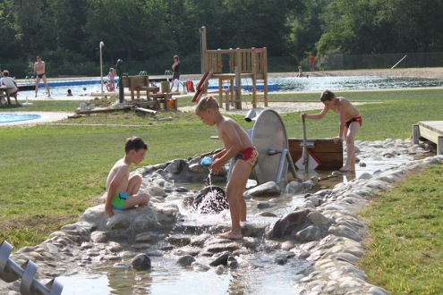 Drei Jungen in Badeanzügen spielen mit Wasser und Steinen in einem flachen Bach in einem Park im Freien, wobei im Hintergrund andere Menschen und ein Spielplatz zu sehen sind.