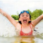 Mädchen im Badeanzug, das mit erhobenen Armen in einem See plantscht, lächelnd mit einer Schwimmbrille auf der Stirn. Im Hintergrund sind Bäume und der Himmel zu sehen.