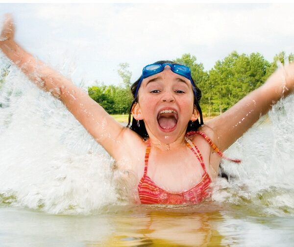 Mädchen im Badeanzug, das mit erhobenen Armen in einem See plantscht, lächelnd mit einer Schwimmbrille auf der Stirn. Im Hintergrund sind Bäume und der Himmel zu sehen.