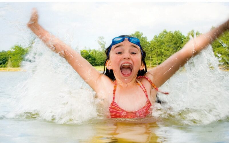 Mädchen im Badeanzug, das mit erhobenen Armen in einem See plantscht, lächelnd mit einer Schwimmbrille auf der Stirn. Im Hintergrund sind Bäume und der Himmel zu sehen.