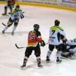 Ein Eishockeyspieler in einem rot-schwarzen Trikot sieht zu, wie der Puck am Torwart vorbei ins Tor geht, während drei Spieler in Weiß verteidigen.