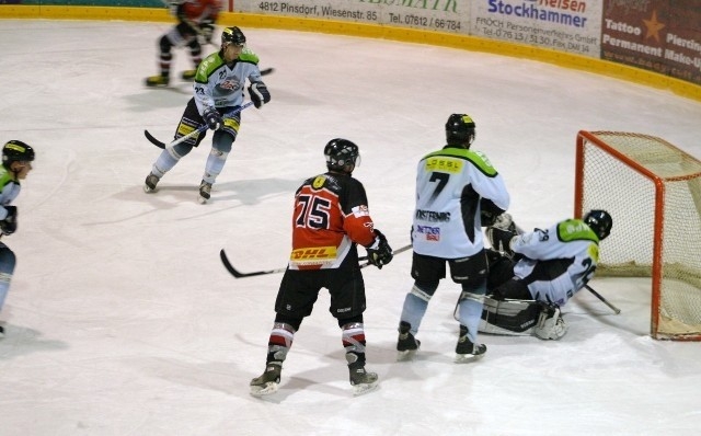 Ein Eishockeyspieler in einem rot-schwarzen Trikot sieht zu, wie der Puck am Torwart vorbei ins Tor geht, während drei Spieler in Weiß verteidigen.