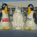 Drei Eislaufhilfsfiguren - zwei Pinguine und ein Eisbär - stehen auf einer Eisbahn und tragen jeweils ein Werbelogo auf ihrer Vorderseite.