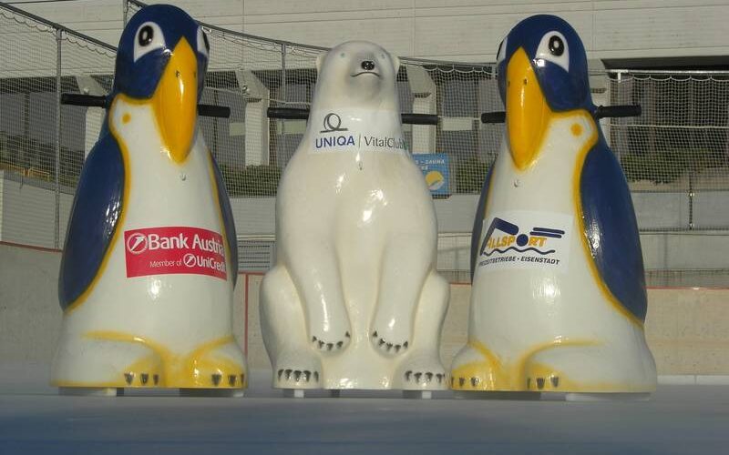 Drei Eislaufhilfsfiguren - zwei Pinguine und ein Eisbär - stehen auf einer Eisbahn und tragen jeweils ein Werbelogo auf ihrer Vorderseite.