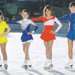 Vier junge Mädchen in farbenfrohen Eiskunstlaufkleidern posieren gemeinsam auf einer Eisbahn im Freien, jede trägt Schlittschuhe und lächelt in die Kamera.