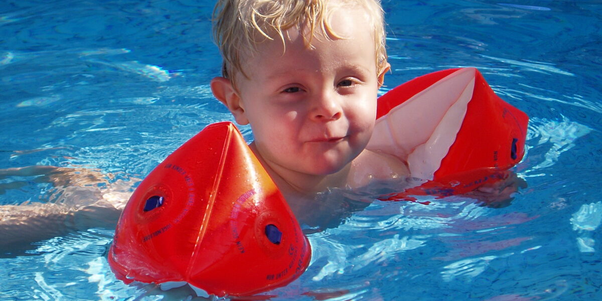 Junges Kind mit blondem Haar, das rote aufblasbare Armbänder trägt, schwimmt in einem Pool mit klarem blauen Wasser.