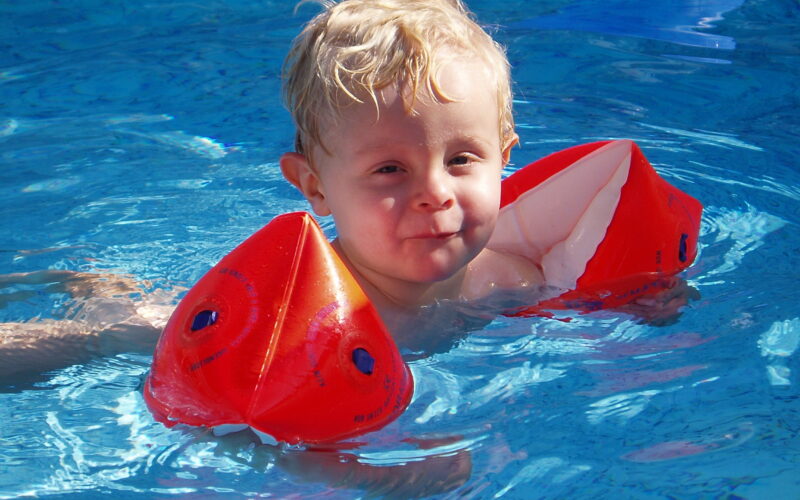 Junges Kind mit blondem Haar, das rote aufblasbare Armbänder trägt, schwimmt in einem Pool mit klarem blauen Wasser.
