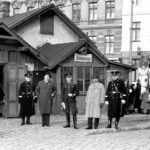Eine Gruppe von uniformierten Männern, einige davon zu Pferd, steht vor einem kleinen Gebäude auf einer Kopfsteinpflasterstraße mit städtischen Gebäuden im Hintergrund.