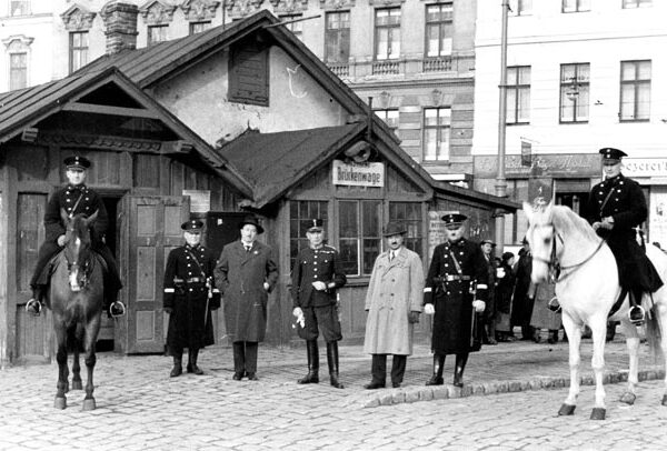 Eine Gruppe von uniformierten Männern, einige davon zu Pferd, steht vor einem kleinen Gebäude auf einer Kopfsteinpflasterstraße mit städtischen Gebäuden im Hintergrund.
