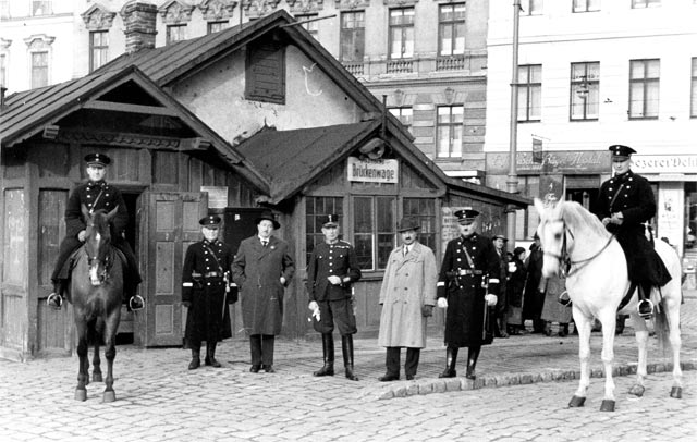 Eine Gruppe von uniformierten Männern, einige davon zu Pferd, steht vor einem kleinen Gebäude auf einer Kopfsteinpflasterstraße mit städtischen Gebäuden im Hintergrund.
