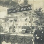 Ein alter Marktstand mit der Aufschrift "Marktamt der Stadt Wien", an dem Flaschen und Gläser ausgestellt sind, umgeben von Menschen, darunter ein Uniformierter im Vordergrund.