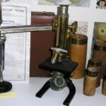 Ein altes Messingmikroskop wird zusammen mit wissenschaftlichen Instrumenten, beschrifteten Behältern und Papieren vor einem Hintergrund aus Postern und Äpfeln ausgestellt.