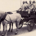 Schwarz-Weiß-Foto einer Pferdekutsche mit uniformierten Männern, die Helme tragen, wahrscheinlich historisches Militär oder Rettungsdienstpersonal.