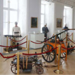 Eine Museumsausstellung zeigt alte Kanonen und Artillerie, umgeben von roten Absperrseilen, mit Schaufensterpuppen in historischen Militäruniformen im Hintergrund.