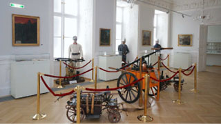 Eine Museumsausstellung zeigt alte Kanonen und Artillerie, umgeben von roten Absperrseilen, mit Schaufensterpuppen in historischen Militäruniformen im Hintergrund.