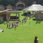 Eine Gruppe von Kindern spaziert an einem sonnigen Tag über eine von Holzgebäuden umgebene Wiese, möglicherweise an einem historischen Ort oder einer Bildungsstätte im Freien.