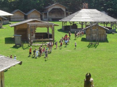 Eine Gruppe von Kindern spaziert an einem sonnigen Tag über eine von Holzgebäuden umgebene Wiese, möglicherweise an einem historischen Ort oder einer Bildungsstätte im Freien.