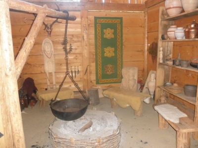 Ein rustikaler Holzraum mit einer zentralen Feuerstelle, einem Hängetopf, Bänken, Regalen mit Töpferwaren und einem grünen Wandteppich an der Wand.