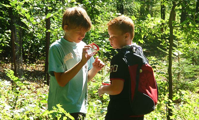 Zwei Jungen stehen in einem sonnenbeschienenen Wald, einer hält eine Lupe in der Hand und untersucht einen Gegenstand, während der andere zusieht und einen Rucksack trägt.