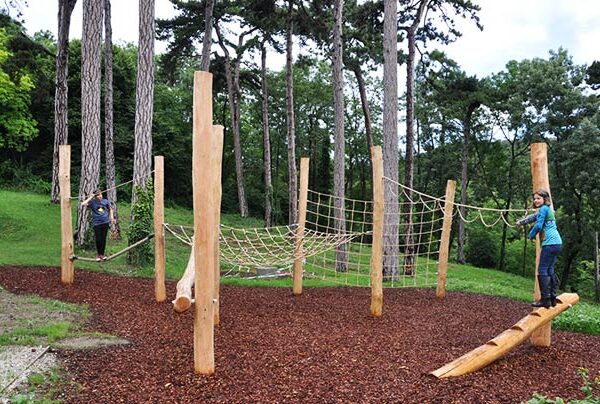 Zwei Personen benutzen ein natürliches Holzspielgerät mit Kletternetzen und Balancierbalken, umgeben von Bäumen und Mulchboden.