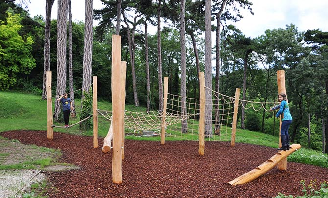Zwei Personen benutzen ein natürliches Holzspielgerät mit Kletternetzen und Balancierbalken, umgeben von Bäumen und Mulchboden.