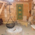 Ein rustikaler Holzraum mit einer zentralen Feuerstelle, einem hängenden Topf, Holzbänken, Regalen mit Töpferwaren und einem grünen Wandteppich an der Wand.