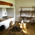 Eine traditionelle Schmiedewerkstatt mit Ambossen, einer Schmiede und verschiedenen Handwerkzeugen, die auf Holzregalen an weißen Wänden hängen.