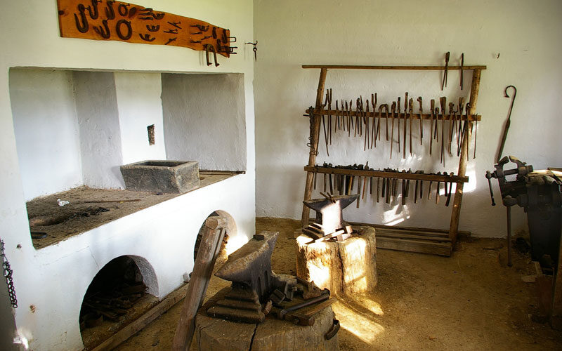 Eine traditionelle Schmiedewerkstatt mit Ambossen, einer Schmiede und verschiedenen Handwerkzeugen, die auf Holzregalen an weißen Wänden hängen.