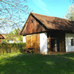 Ein rustikales Holzhaus mit schrägem Dach steht in einem grasbewachsenen Hof mit Bäumen und Sonnenlicht, neben einer Hecke und einer weiteren Holzkonstruktion im Hintergrund.