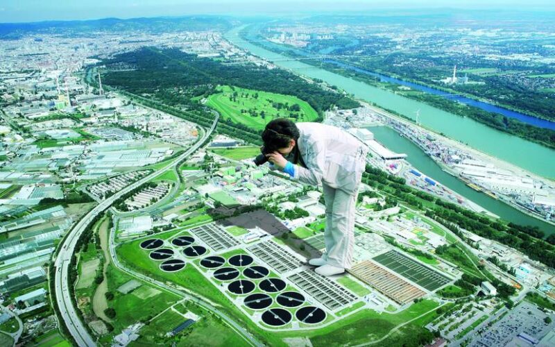 Ein digital verfremdetes Bild zeigt eine übergroße Person mit einer Kamera, die auf einem Luftbild einer Stadt mit einem Fluss, Autobahnen und Industriegebäuden steht.