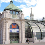 In Wien steht das Schmetterlinghaus aus Glas und Stein mit kunstvollen Statuen und Rundbogenfenstern; eine Person geht am Eingang vorbei.