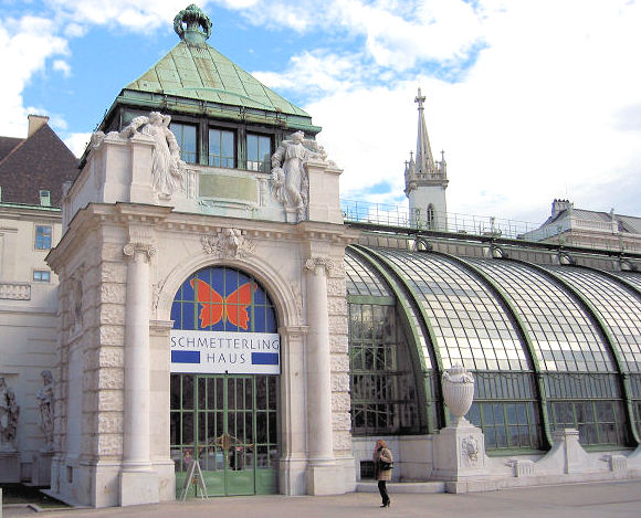 In Wien steht das Schmetterlinghaus aus Glas und Stein mit kunstvollen Statuen und Rundbogenfenstern; eine Person geht am Eingang vorbei.