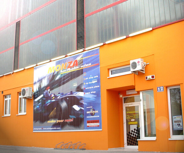 Leuchtend orangefarbenes Gebäude mit mehreren Fenstern, einer Glastür und einem großen Werbeschild der Monza Kartbahn Wien, auf dem ein fahrender Rennwagen abgebildet ist.